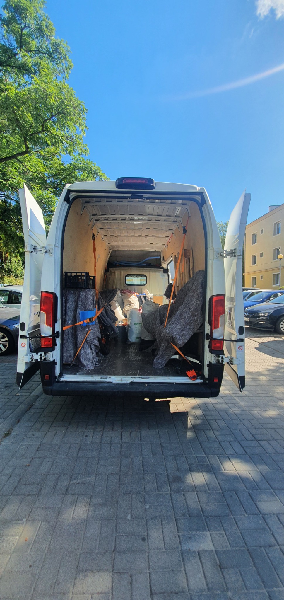 Wnętrze białego busa z otwartymi drzwiami, zabezpieczony ładunek kocami i pasami transportowymi, widok z tyłu na zaparkowane samochody i budynek w tle.