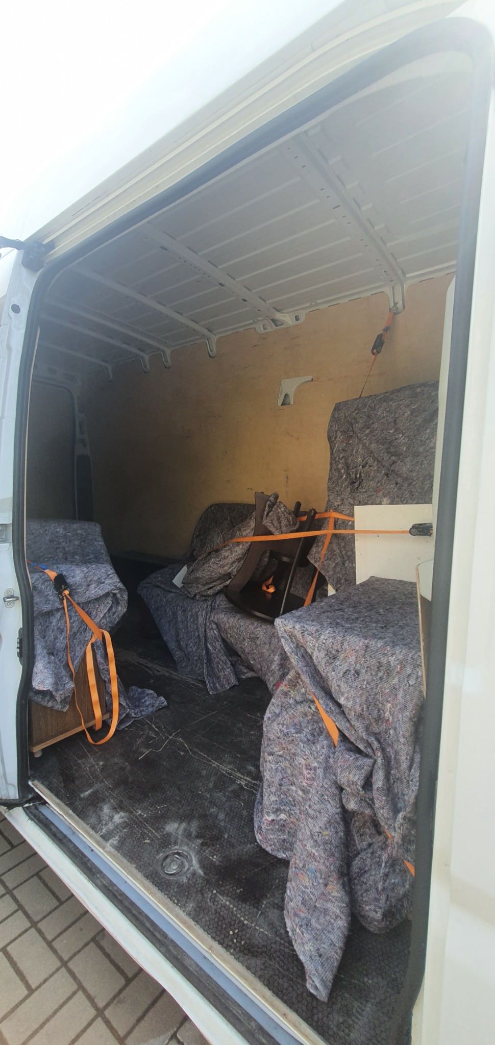 Wnętrze białego busa z otwartymi drzwiami, zabezpieczone meble pod kocami transportowymi i spięte pomarańczowymi pasami.