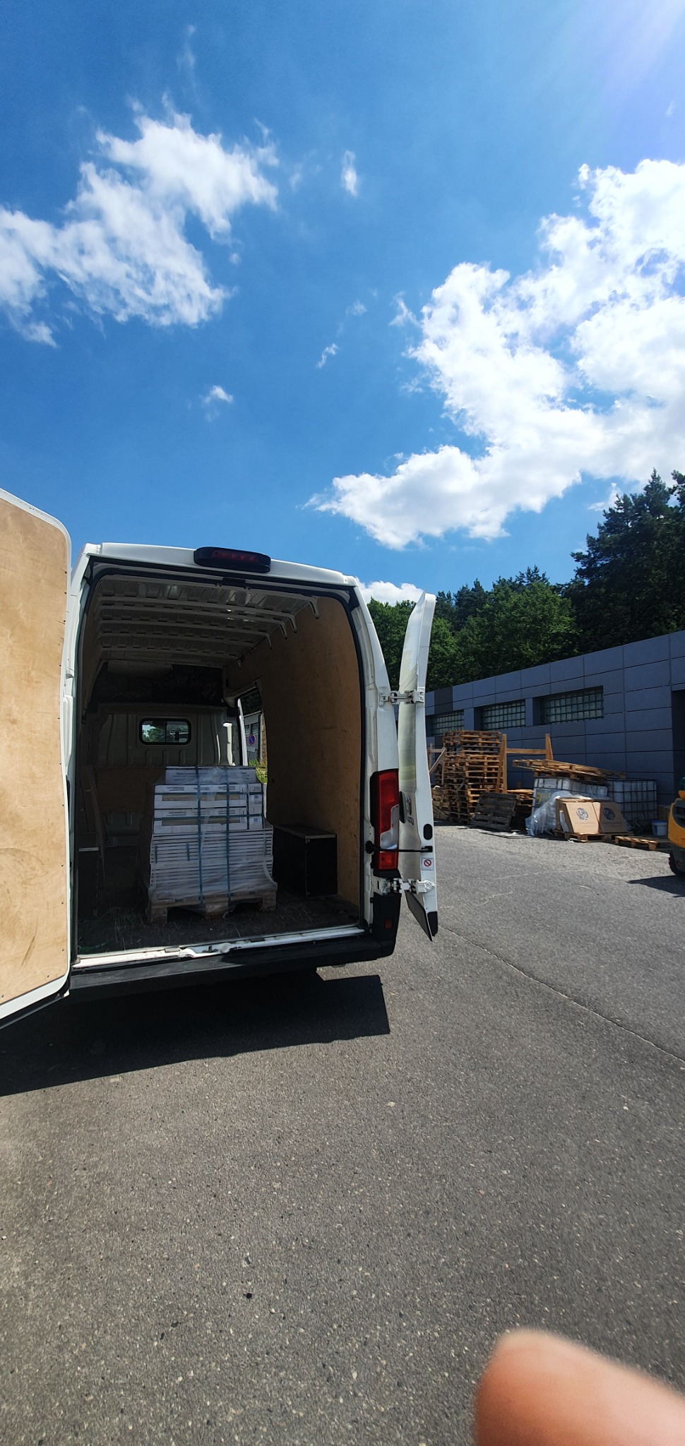 Wnętrze białego busa z otwartymi drzwiami, załadowanego paletą z pakietami płyt w folii, widok z zewnątrz na parking i budynek przemysłowy w tle pod błękitnym niebem z chmurami.