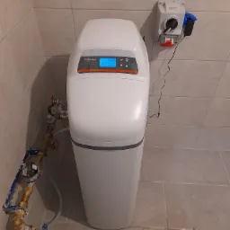 Biały zmiękczacz wody Viessmann Aquastilla 25 z podłączeniem hydraulicznym i elektrycznym w pomieszczeniu z beżowymi płytkami.