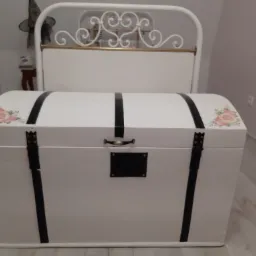 Biała skrzynia na pościel z czarnymi paskami i kwiatowymi zdobieniami, ustawiona na podłodze w pokoju, z zagłówkiem łóżka w tle.