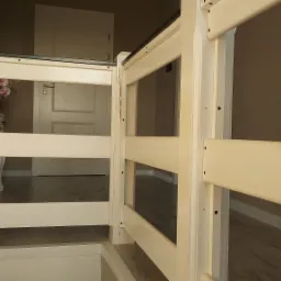 Biała drewniana balustrada antresoli z widocznym fragmentem schodów, przez szczeliny widać stolik z kwiatami i drzwi w tle.