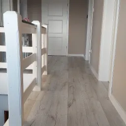 Jasne panele podłogowe w przedpokoju z białymi drzwiami i białą balustradą schodów, widok z góry schodów pokrytych ciemną wykładziną.