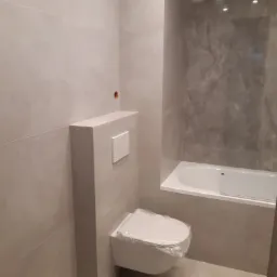 Wykończona łazienka z podwieszaną toaletą z przyciskiem spłukującym, obudową i płytkami w odcieniach szarości oraz wanną z szarą ścianą imitującą marmur.