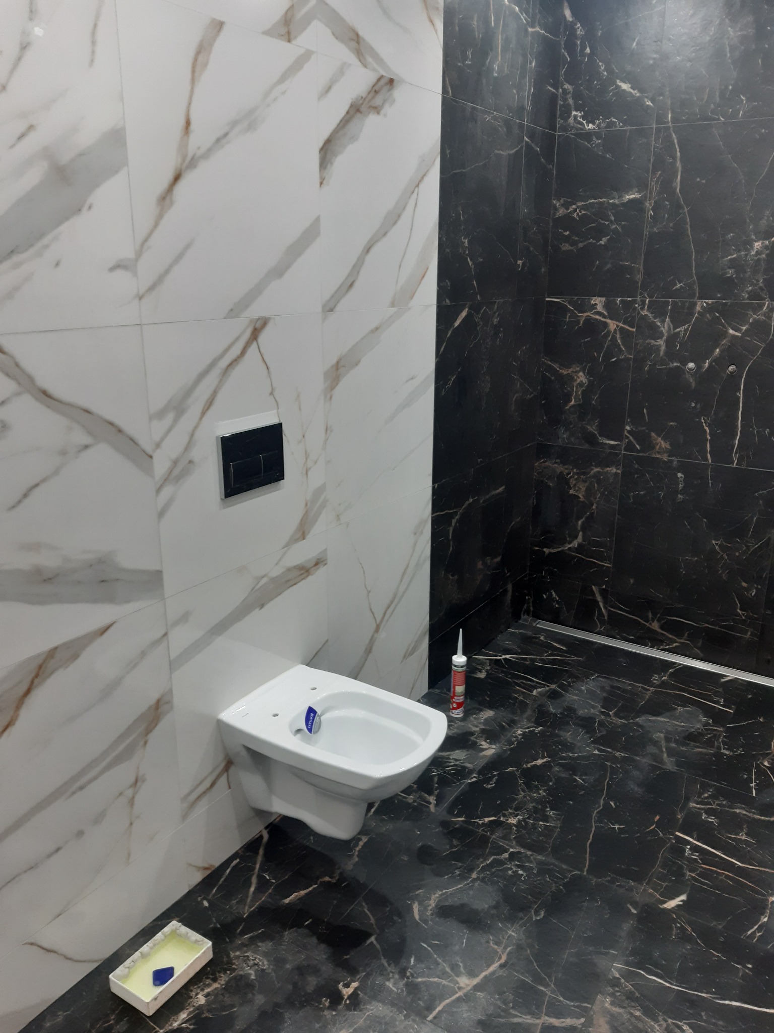 Montaż podwieszanej miski WC w łazience z czarno-białymi marmurowymi płytkami, widoczny aplikator silikonu i pojemnik z fugą.