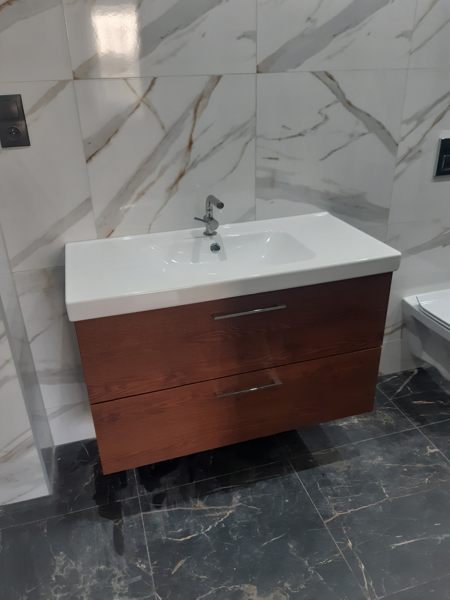 Nowoczesna umywalka z drewnianą szafką z dwoma szufladami i chromowaną armaturą na tle marmurowych płytek ściennych i podłogowych w łazience.