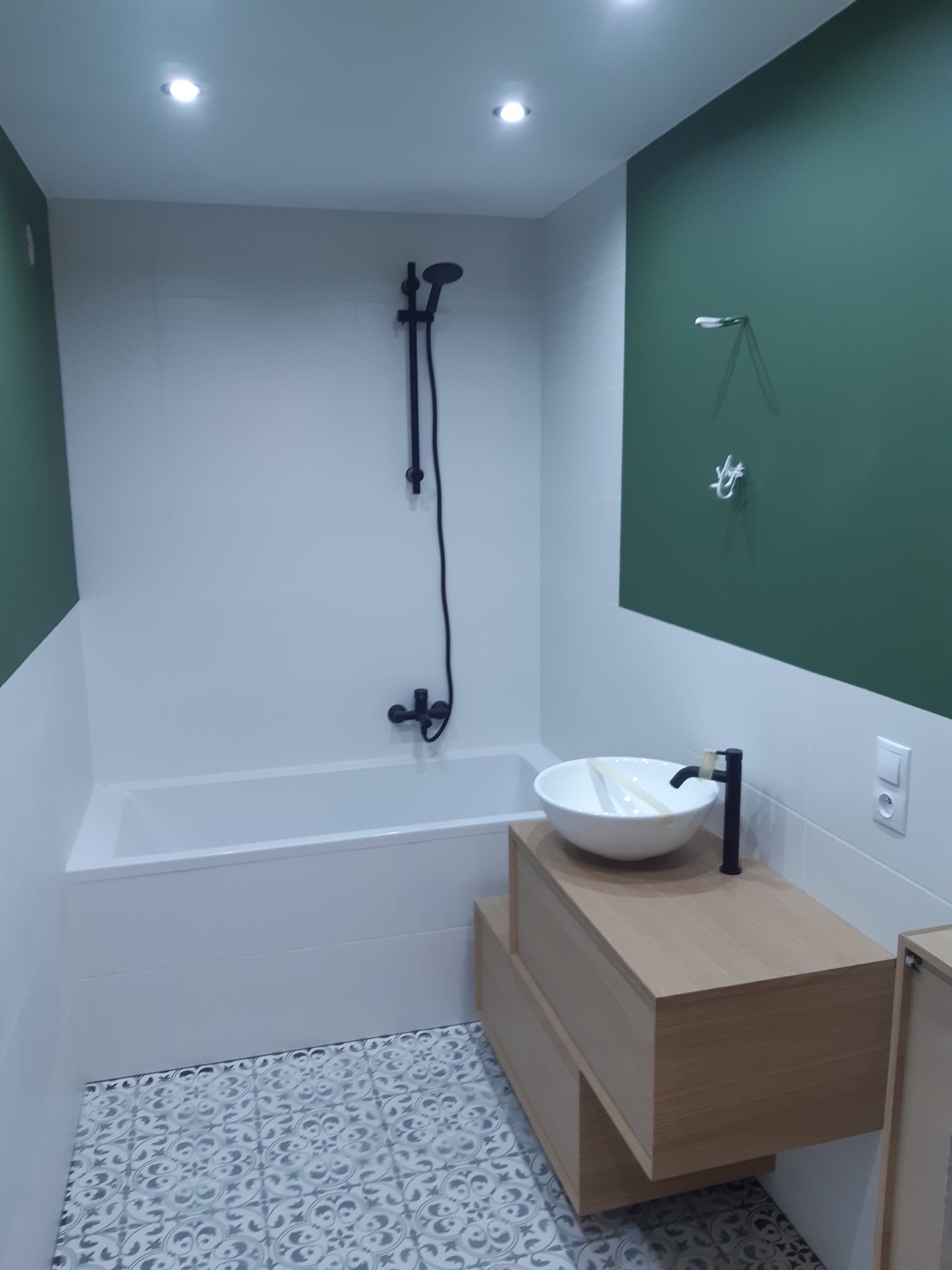 Nowoczesna łazienka z białą wanną, umywalką nablatową na drewnianej szafce, czarną armaturą i zielonym akcentem kolorystycznym na ścianie.