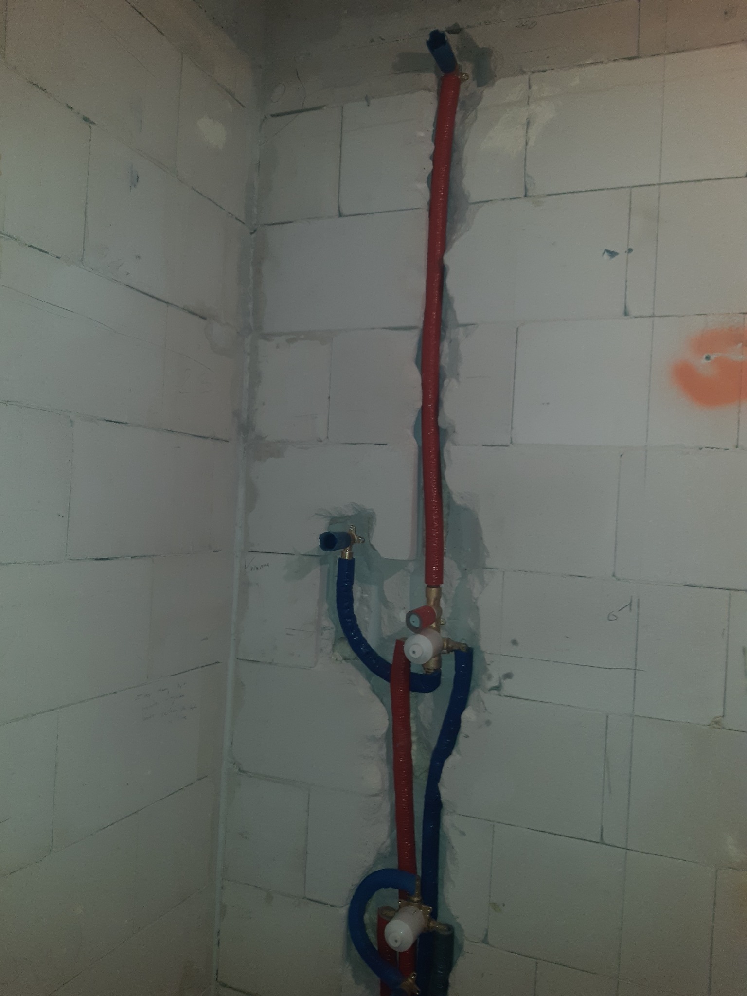 Surowe ściany z betonu komórkowego w rogu pomieszczenia z poprowadzonymi rurami instalacji wodnej w kolorach czerwonym i niebieskim, widoczne elementy armatury.