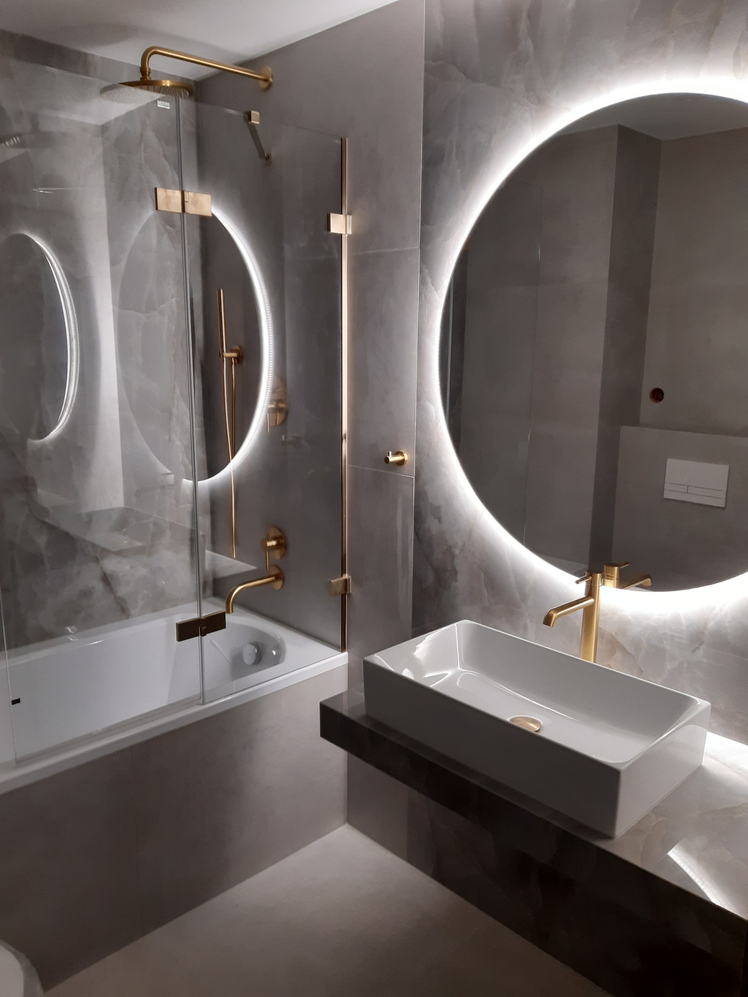 Nowoczesna łazienka z szarą marmurową okładziną, owalnymi lustrami z podświetleniem LED, złotą armaturą i prostokątną umywalką nablatową.