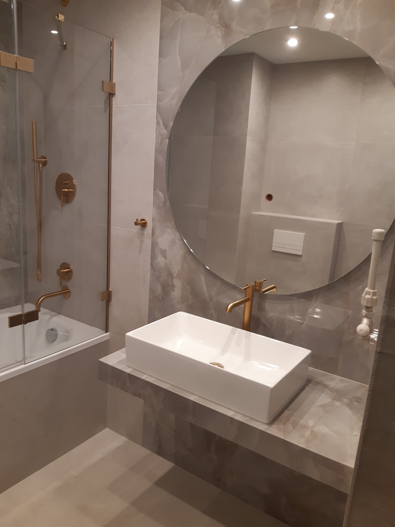 Nowoczesna łazienka z umywalką nablatową, baterią i armaturą w kolorze złotym, okrągłe lustro oraz kabina prysznicowa z elementami w kolorze złotym, szare płytki imitujące marmur na ścianach...