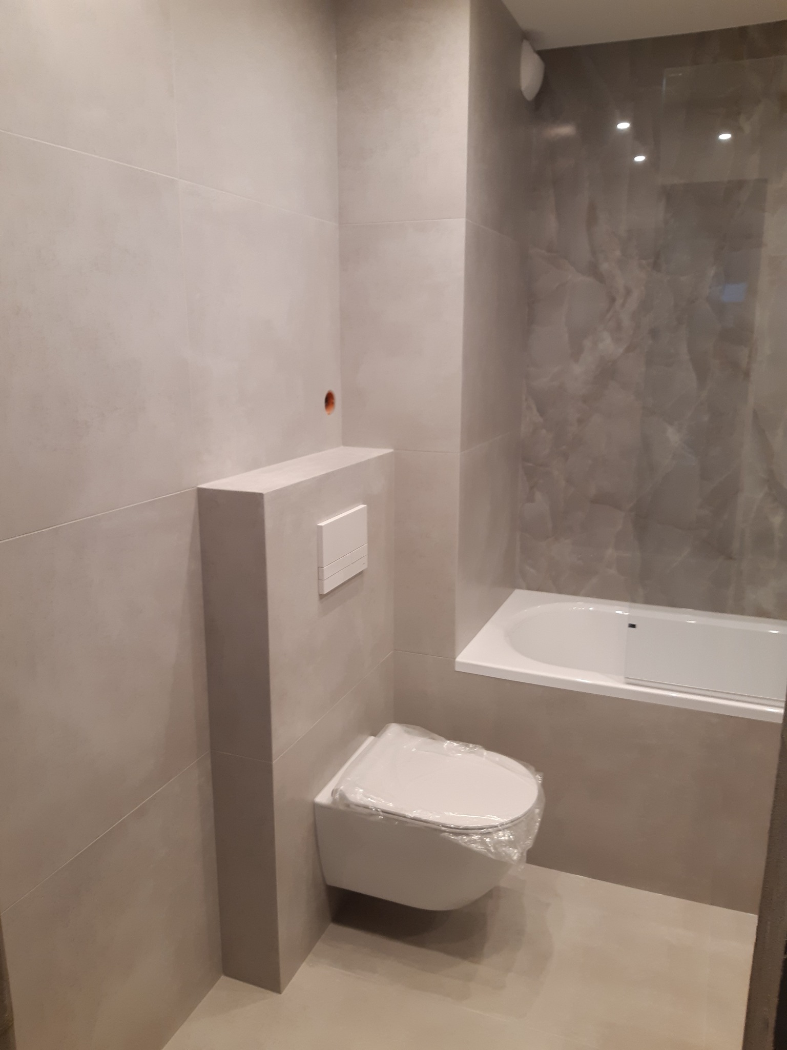 Wykończona łazienka z podwieszaną toaletą z przyciskiem spłukującym, obudową i płytkami w odcieniach szarości oraz wanną z szarą ścianą imitującą marmur.