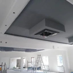 Wnętrze w trakcie remontu: pomalowany na szaro sufit z ozdobnymi wnękami i przygotowanymi otworami na oświetlenie, widoczne rusztowania i drabina.