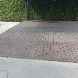 Zadaszony taras z desek kompozytowych, otoczony tujami i wykończony szarą kostką brukową.