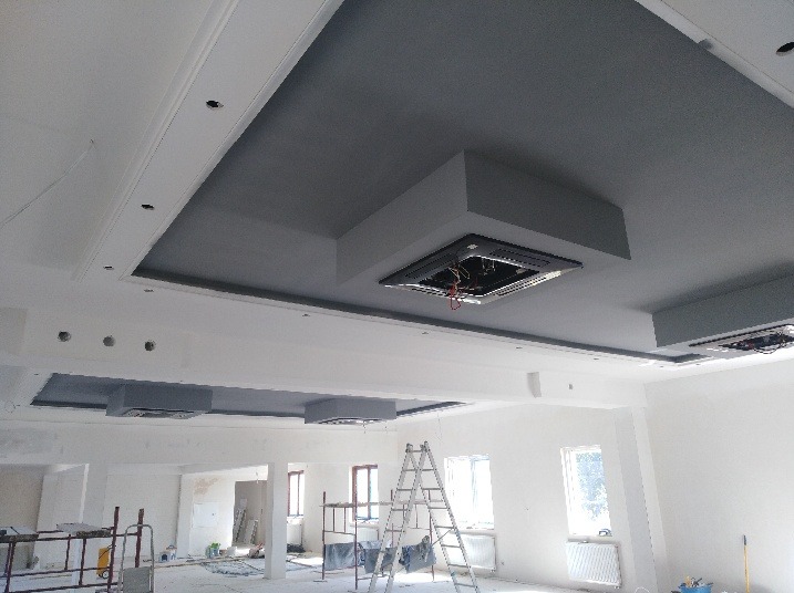 Wnętrze w trakcie remontu: pomalowany na szaro sufit z ozdobnymi wnękami i przygotowanymi otworami na oświetlenie, widoczne rusztowania i drabina.