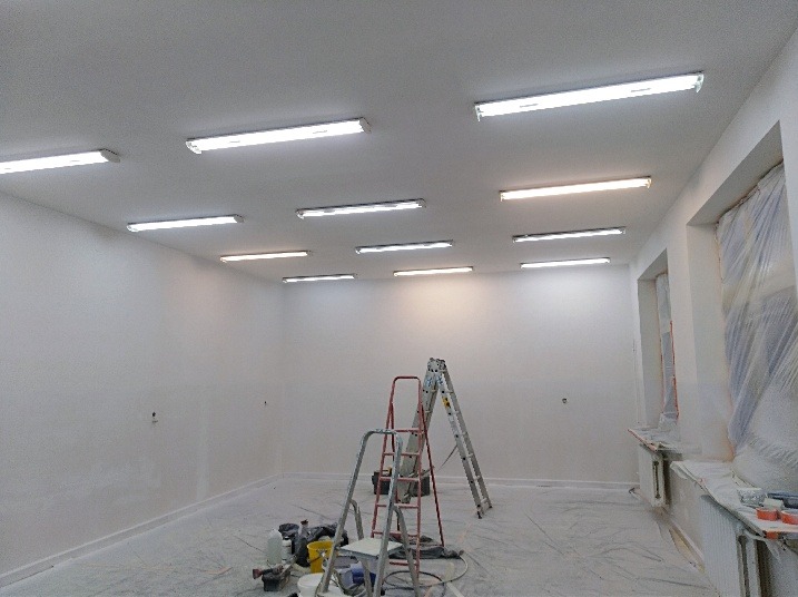 Wnętrze pomieszczenia po świeżym pomalowaniu ścian na biało, widoczne dwie drabiny, zakryte folią okna, liczne lampy sufitowe i akcesoria malarskie na podłodze.