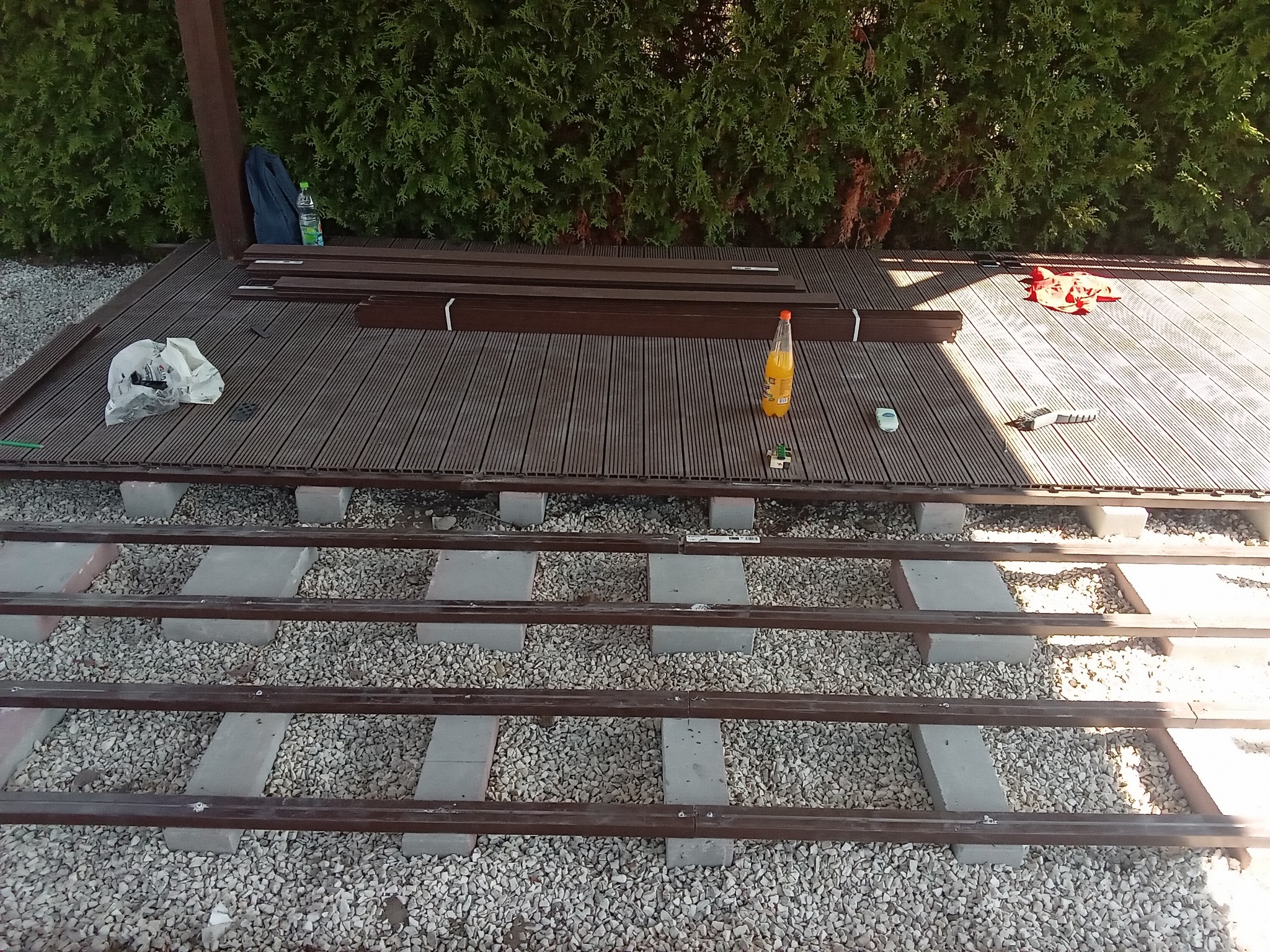 Konstrukcja pod taras z kompozytu na betonowych bloczkach i żwirowym podłożu; deski kompozytowe ułożone częściowo, narzędzia i napoje obok.