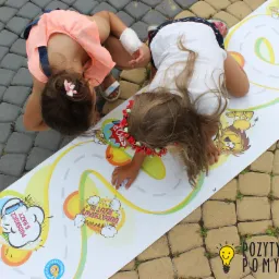 Dwie dziewczynki bawią się na chodniku na rozłożonej planszy do gry z motywami kreskówkowymi, jedna z opaską kwiatową na głowie, druga z opatrunkiem na dłoni.