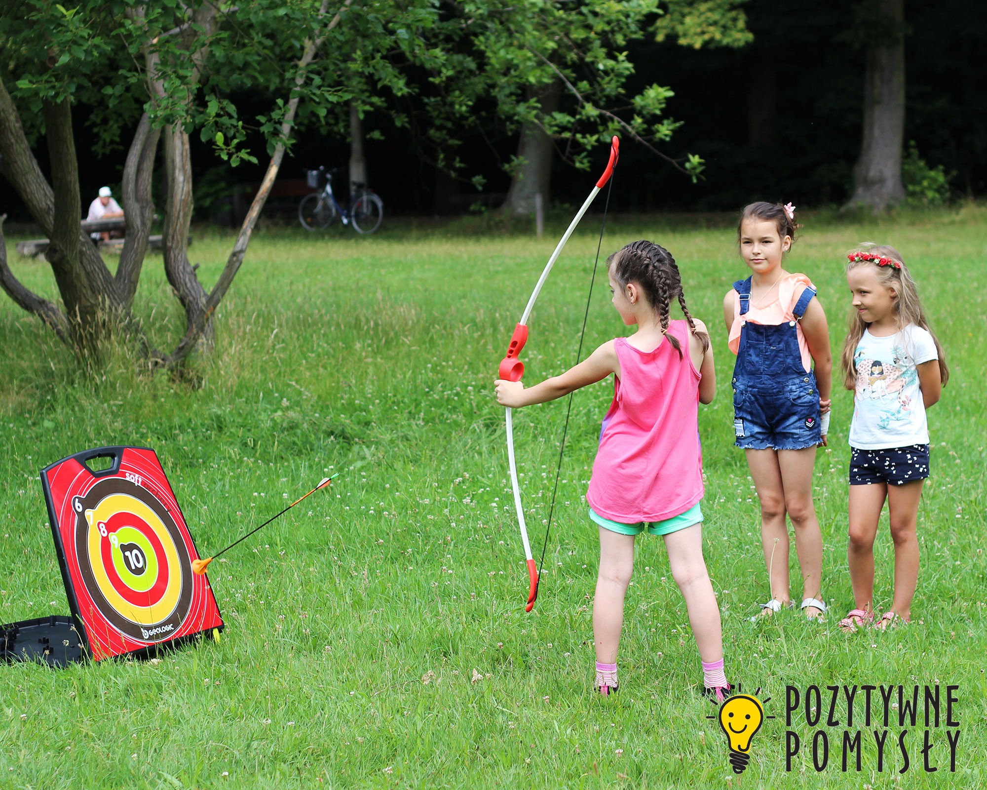 Dziewczynka z warkoczami naciąga łuk, celując do tarczy z numerami na trawie, dwie inne dziewczynki obserwują, w tle zieleń parku i spacerujący ludzie.