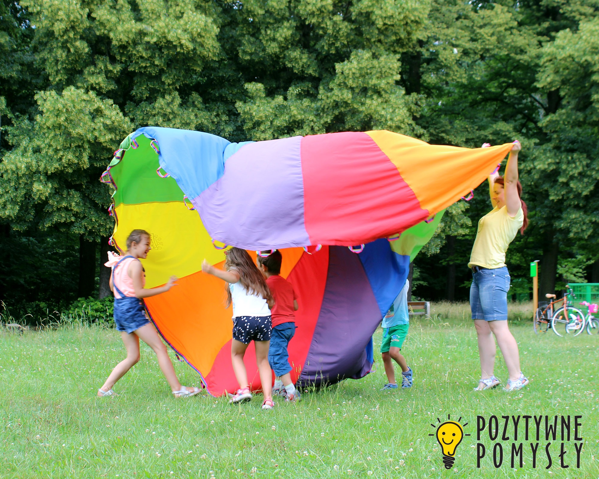 Grupa dzieci i animatorka bawi się kolorową chustą animacyjną na trawie w parku, w tle widoczne drzewa i rowery.