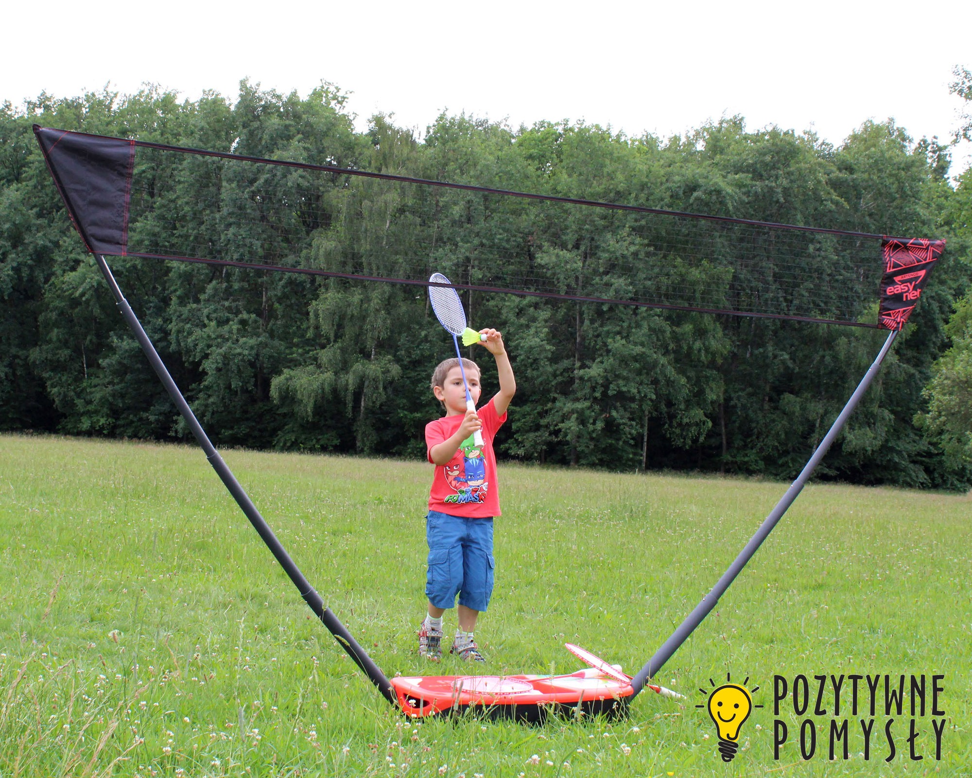 Chłopiec w czerwonej koszulce z bajkowymi postaciami, trzymający rakietę do badmintona i lotkę na tle rozstawionej siatki do badmintona na zielonej łące.