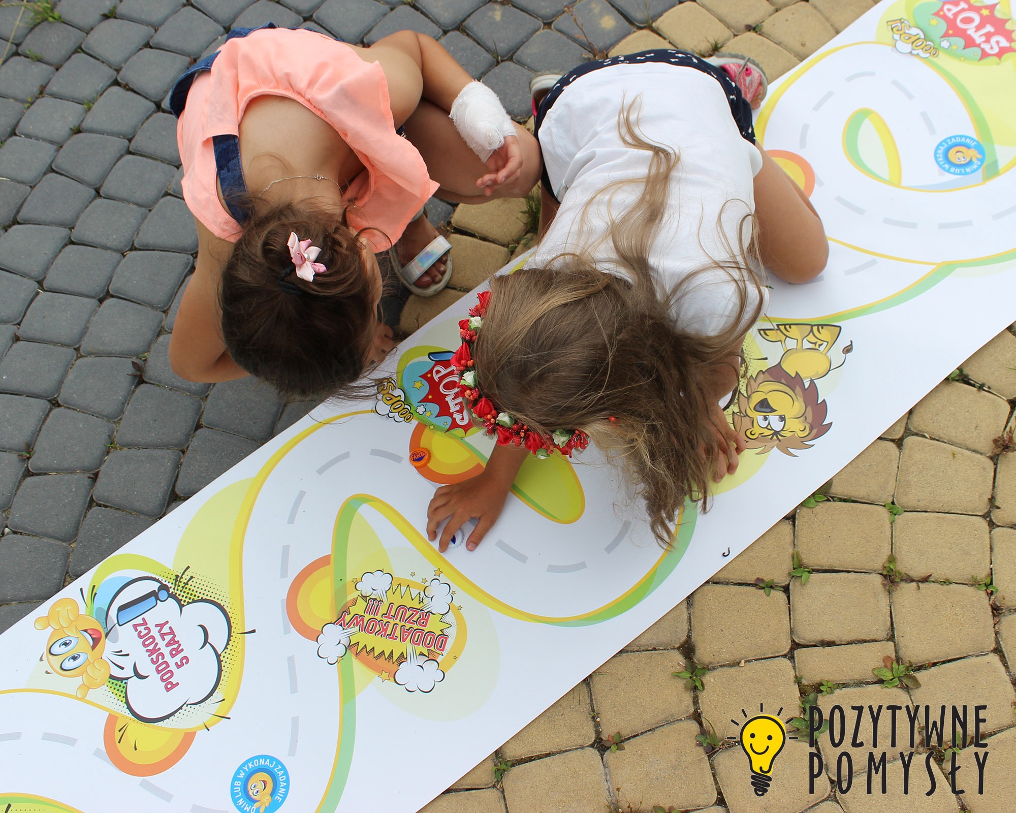 Dwie dziewczynki bawią się na chodniku na rozłożonej planszy do gry z motywami kreskówkowymi, jedna z opaską kwiatową na głowie, druga z opatrunkiem na dłoni.