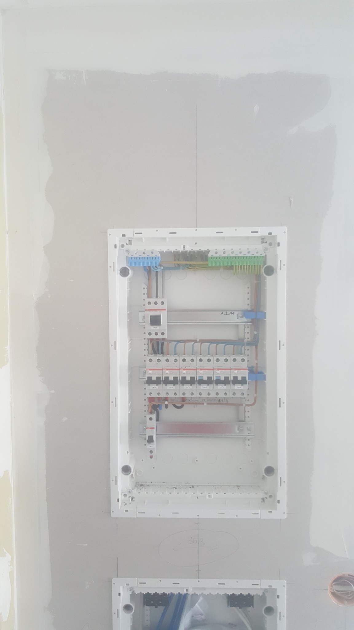 Rozdzielnica elektryczna w trakcie montażu w ścianie, widoczne przewody, bezpieczniki i listwy zaciskowe, ściana po szpachlowaniu, w tle widoczny otwór na kolejną rozdzielnicę.