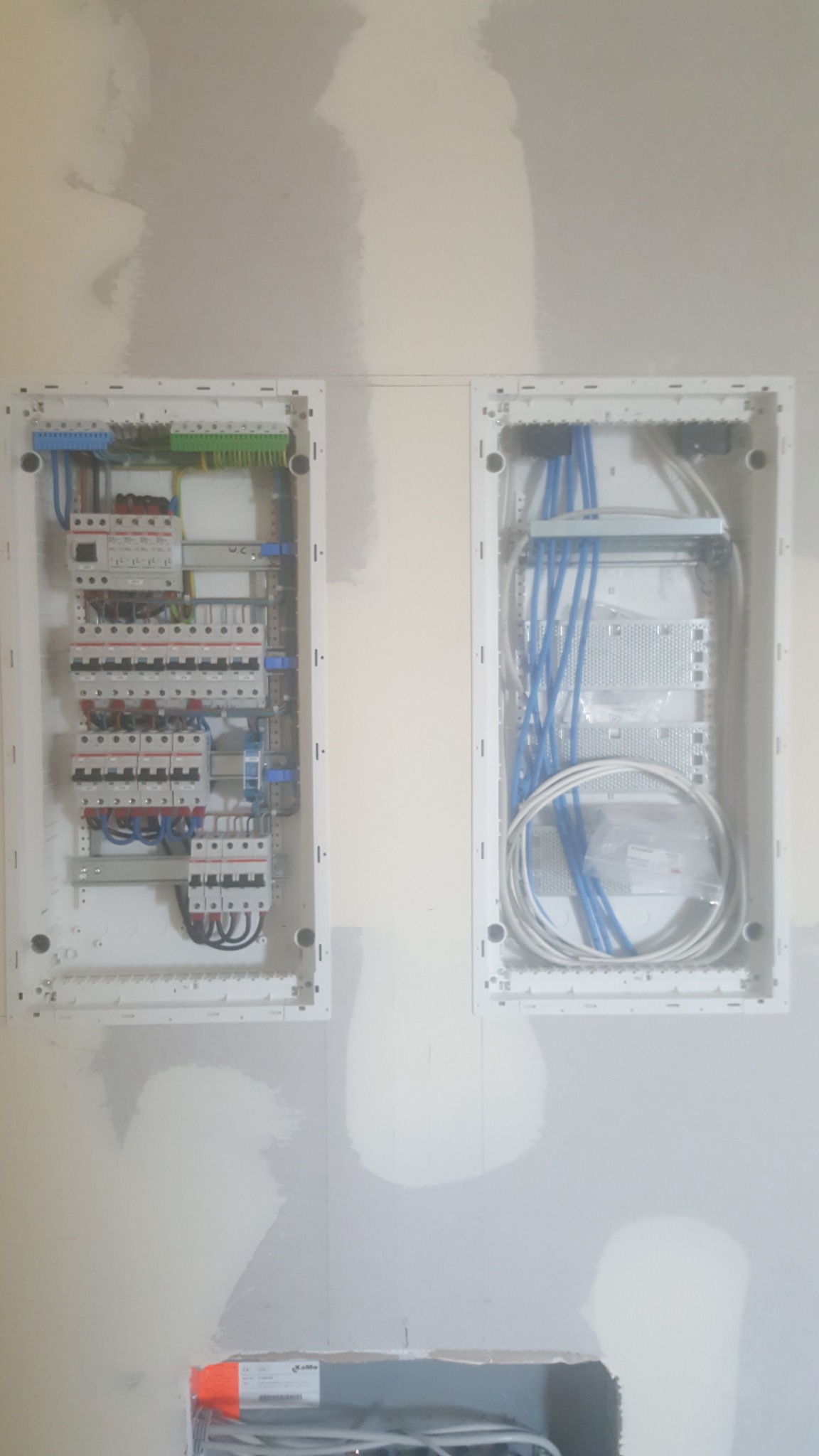 Dwie podtynkowe rozdzielnice elektryczne w trakcie montażu w surowej ścianie, z widocznymi przewodami i elementami zabezpieczeń, w tym jedna z niebieskimi przewodami sieciowymi.