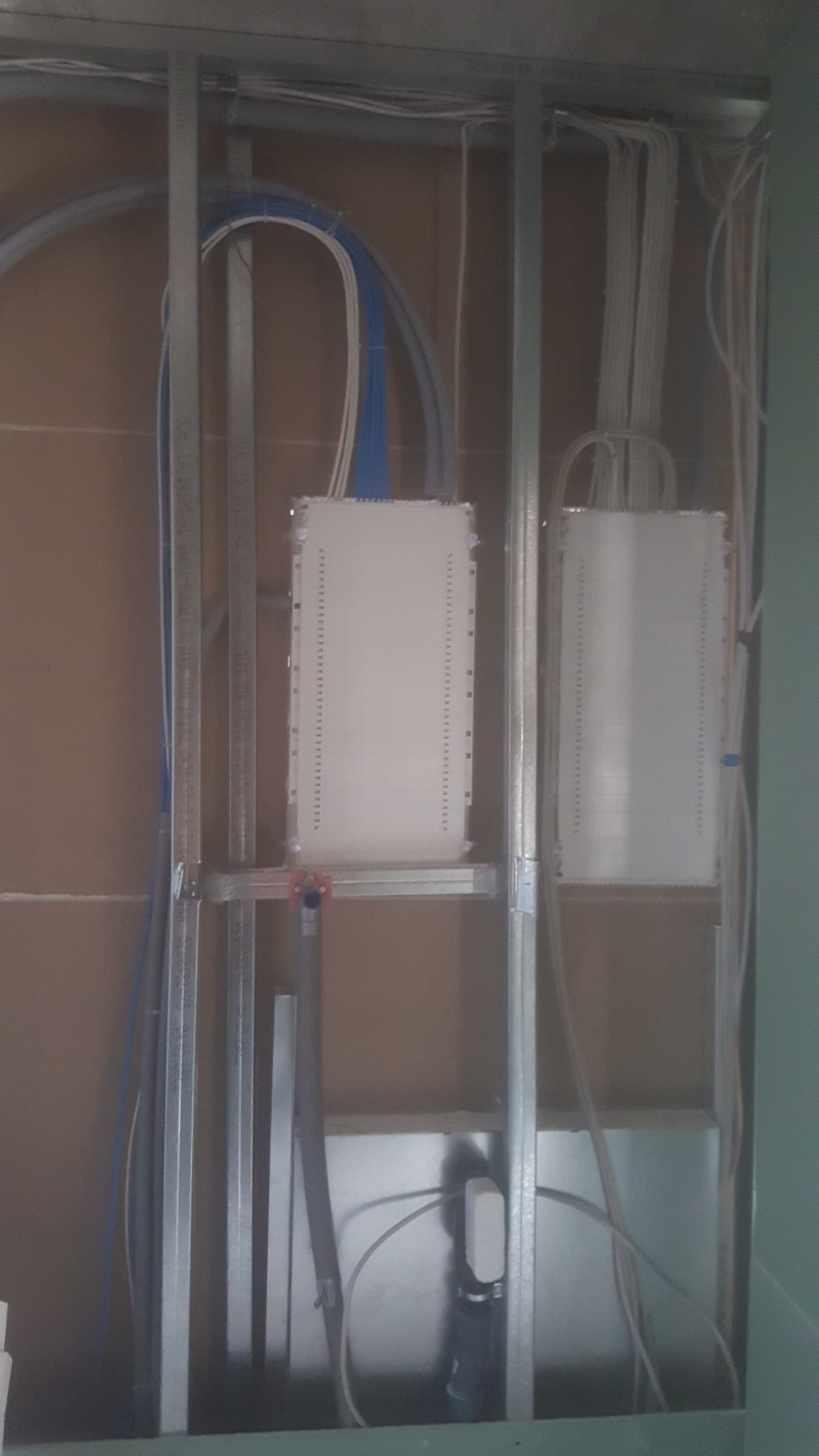 Dwa białe panele krosownicze zamontowane w metalowej konstrukcji szkieletowej, z widocznymi niebieskimi i białymi kablami oraz rurami instalacyjnymi.