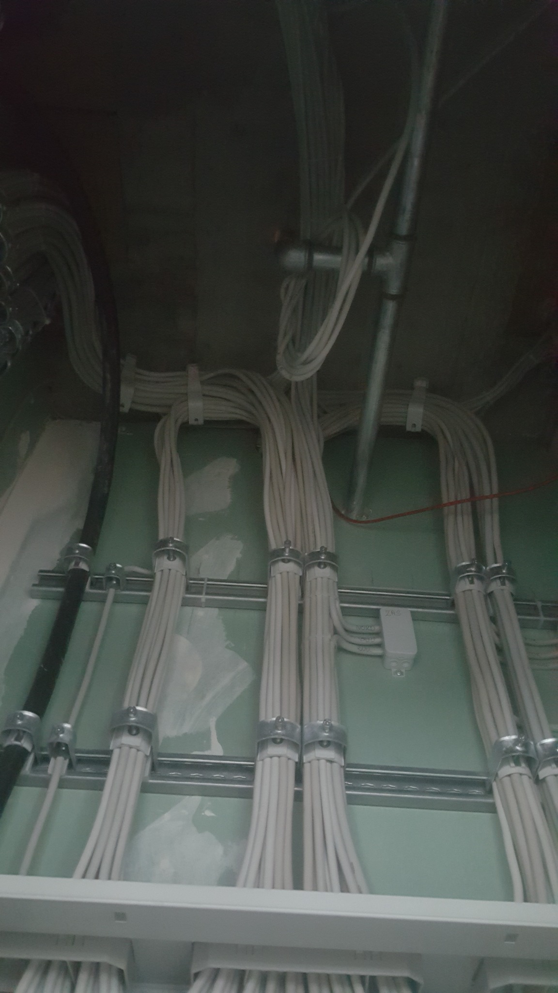 Wiązki kabli elektrycznych przymocowane metalowymi uchwytami do szyn montażowych na zielonej ścianie, widoczne także rury instalacyjne i puszka elektryczna.