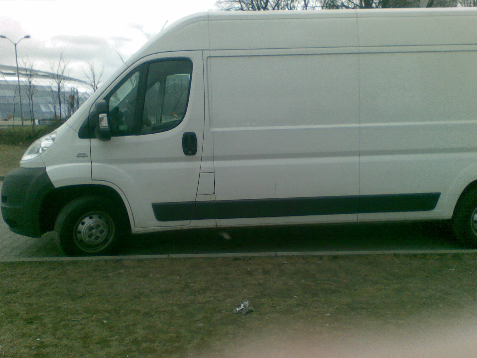 Biały samochód dostawczy Fiat Ducato zaparkowany na poboczu, widok z boku, z fragmentem trawnika i chodnika w kadrze.