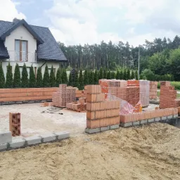 Budowa domu jednorodzinnego w trakcie wznoszenia ścian z czerwonej cegły, widoczne fundamenty, palety z cegłami i gotowy dom w tle.