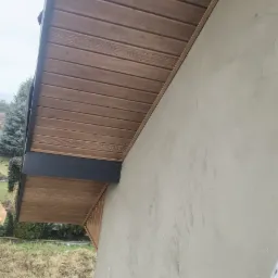 Drewniana podbitka dachowa z czarnym wykończeniem, widok z dołu na tle szarej ściany budynku i trawnika.