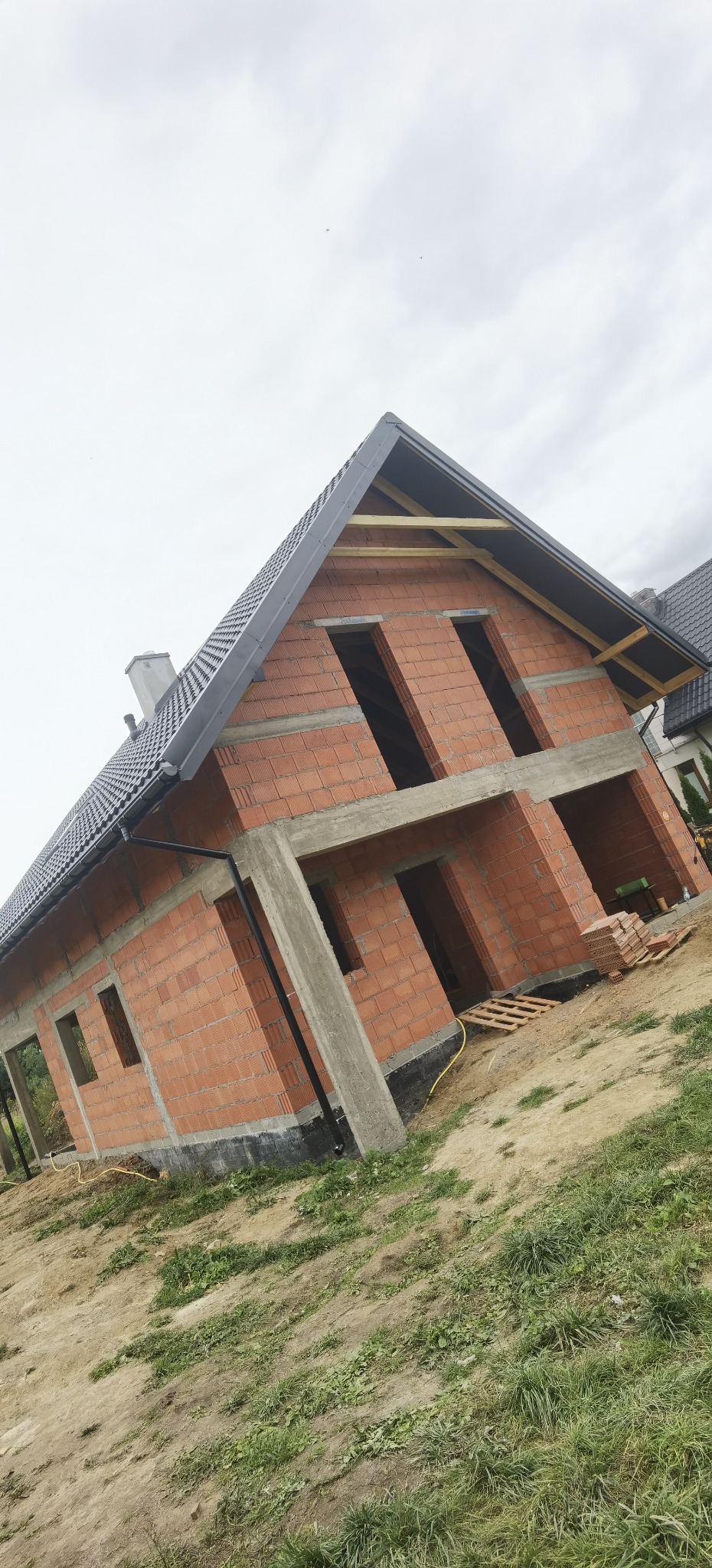 Surowy stan domu murowanego z czerwonej cegły z widocznym betonowym szkieletem, czarnym dachem i drewnianą konstrukcją dachu, na tle pochmurnego nieba.