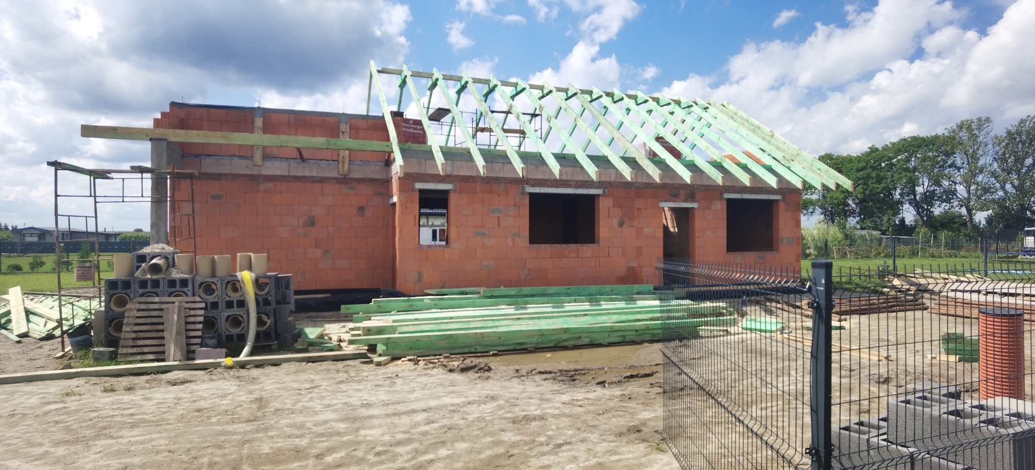 Budowa domu jednorodzinnego z czerwonej cegły, widoczna konstrukcja dachu z zielonego drewna, na pierwszym planie materiały budowlane i ogrodzenie panelowe.