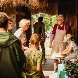 Rzemieślnik w fartuchu uczy dziewczynkę toczenia na kole garncarskim, otoczony grupą obserwujących osób w drewnianej chacie z trzcinowym dachem.