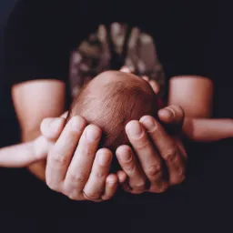 Dłonie dorosłego, z widoczną obrączką, delikatnie obejmujące główkę noworodka na ciemnym tle.