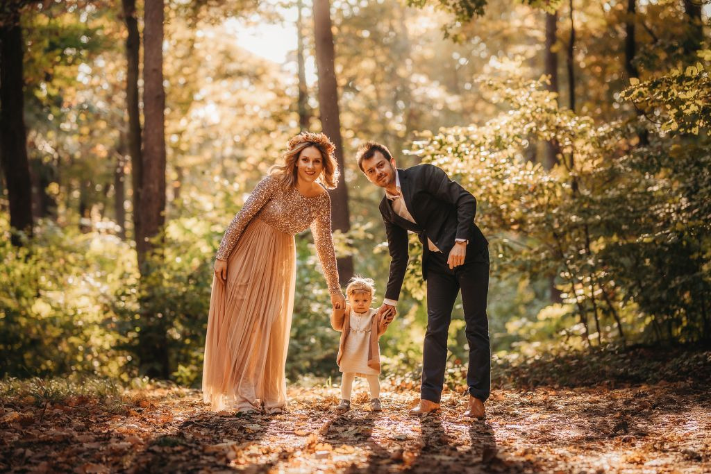 Rodzina z małym dzieckiem pozuje w jesiennym lesie, kobieta w długiej, błyszczącej sukni, mężczyzna w garniturze, dziecko w jasnym ubranku, ciepłe światło słoneczne przebija się przez drzewa...