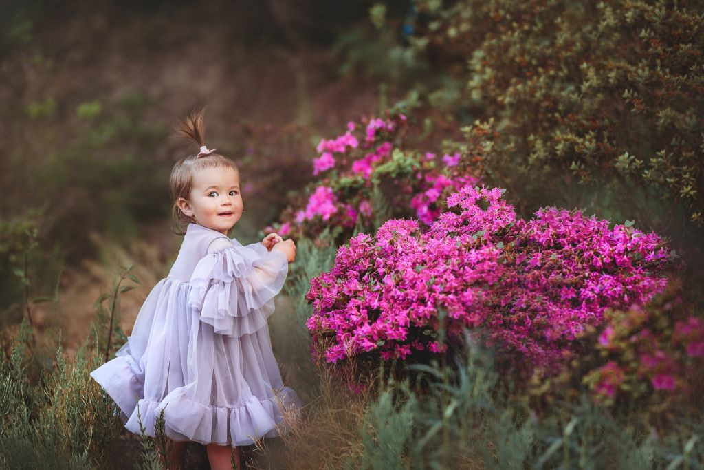 Uśmiechnięta dziewczynka w tiulowej sukience z falbanami, pozująca obok kwitnących różowych azalii w ogrodzie, z włosami upiętymi w mały kucyk.