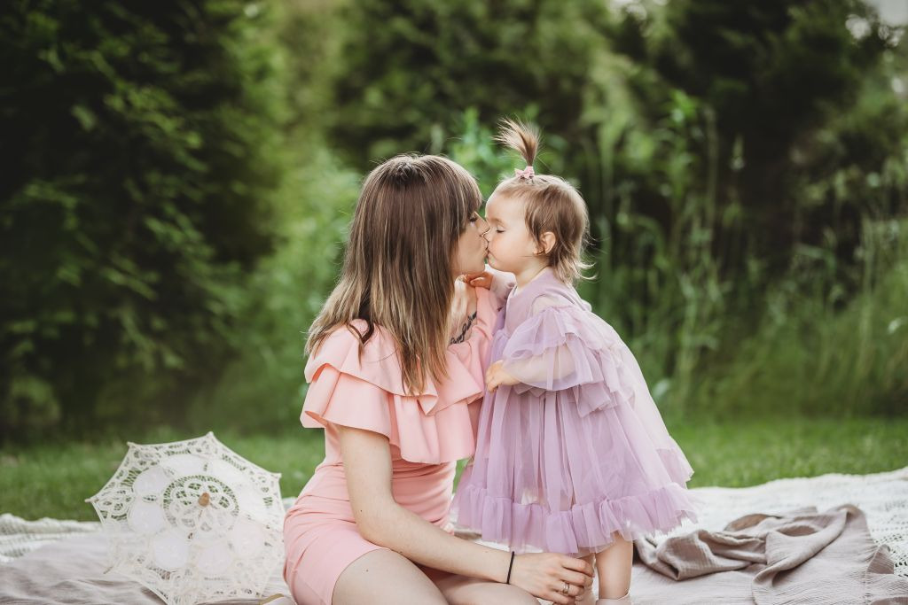 Mama w pudrowo-różowej sukience całuje w usta córeczkę w liliowej sukience na tle zieleni, na kocu leży biały, koronkowy parasol.