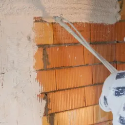 Nakładanie tynku maszynowego na ścianę z czerwonej cegły, widoczny fragment pracownika w białym kombinezonie i okno zabezpieczone folią.
