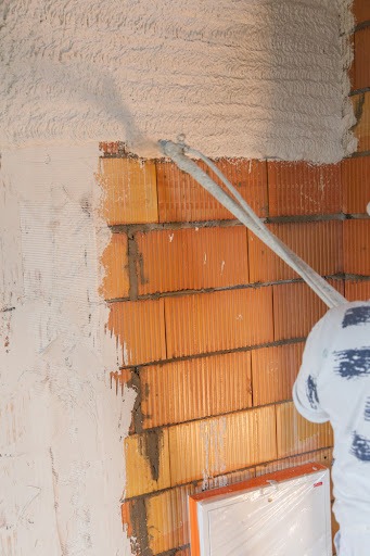 Nakładanie tynku maszynowego na ścianę z czerwonej cegły, widoczny fragment pracownika w białym kombinezonie i okno zabezpieczone folią.