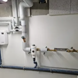 Węzeł instalacji wodnej z filtrem Conel, zaworami kulowymi i rurami z tworzywa, zamontowany na białej ścianie w nowym budynku, widoczne elementy instalacji pod sufitem.