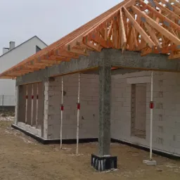 Surowy stan domu w trakcie budowy: ściany z białych bloczk&oacute;w, betonowy wieniec, drewniana konstrukcja dachu z widocznymi stalowymi łącznikami, podpory pionowe z regulacją wysokości.