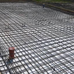 Zbrojenie fundamentu ułożone na izolacji, widoczne rury kanalizacyjne wystające ponad siatkę, drewniany szalunek po obrysie.