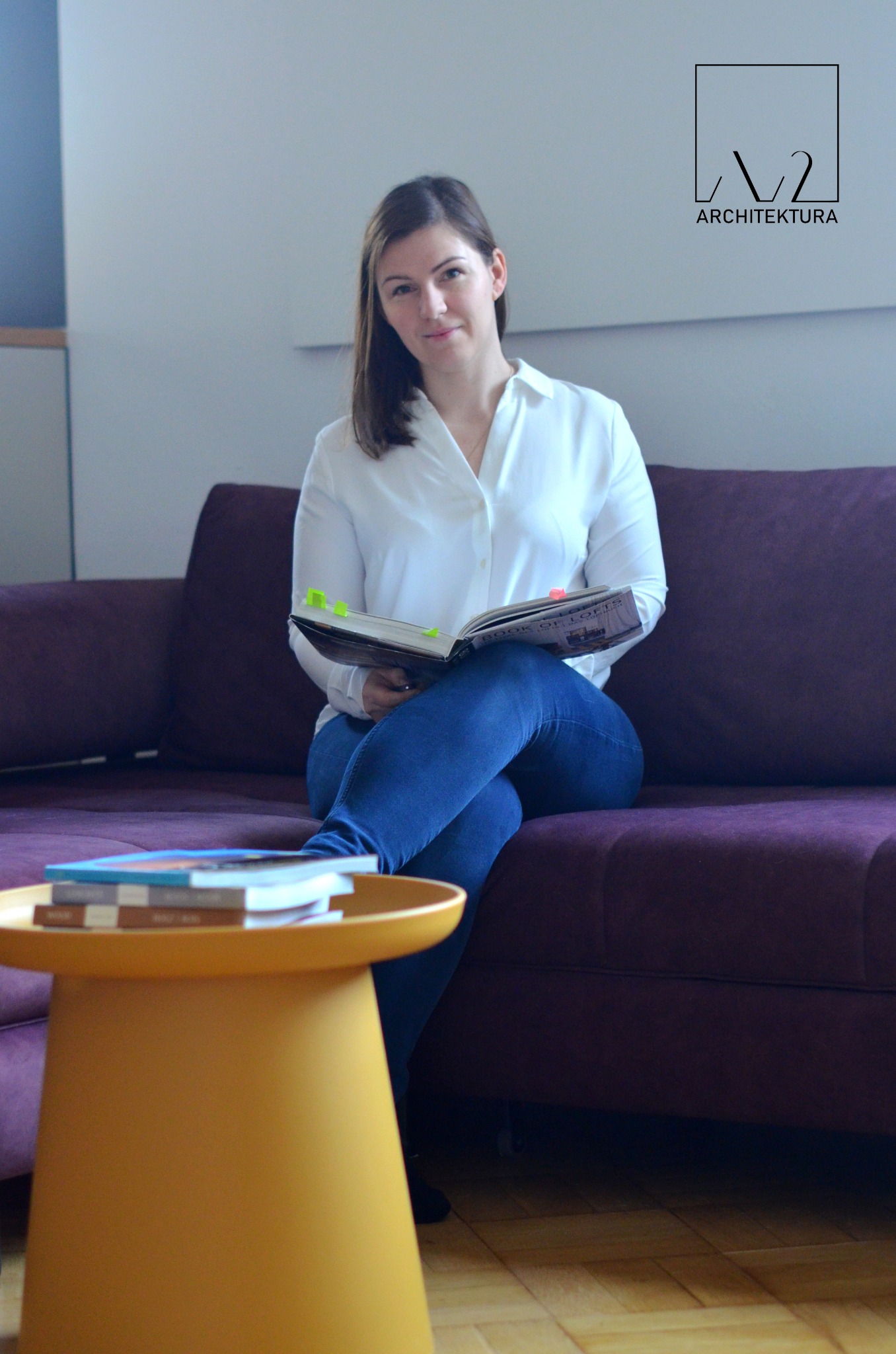 Portret kobiety architekta siedzącej na fioletowej sofie z otwartą książką 'Book of Lofts' z zakładkami, obok żółty stolik z książkami, w tle logo pracowni architektonicznej.
