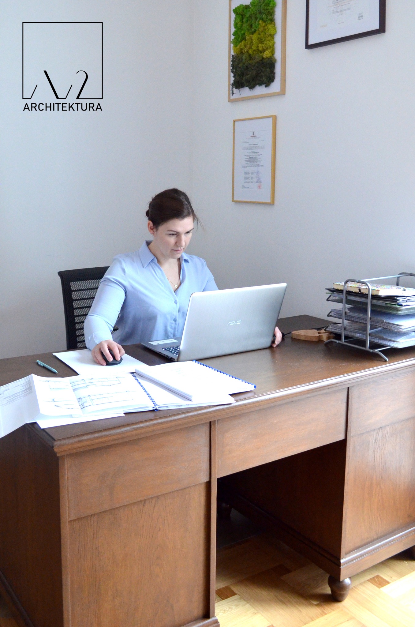 Architektka Anna Korcz-Zając pracuje przy biurku z laptopem, przeglądając plany architektoniczne. Na ścianie wisi logo firmy architektonicznej oraz certyfikaty.