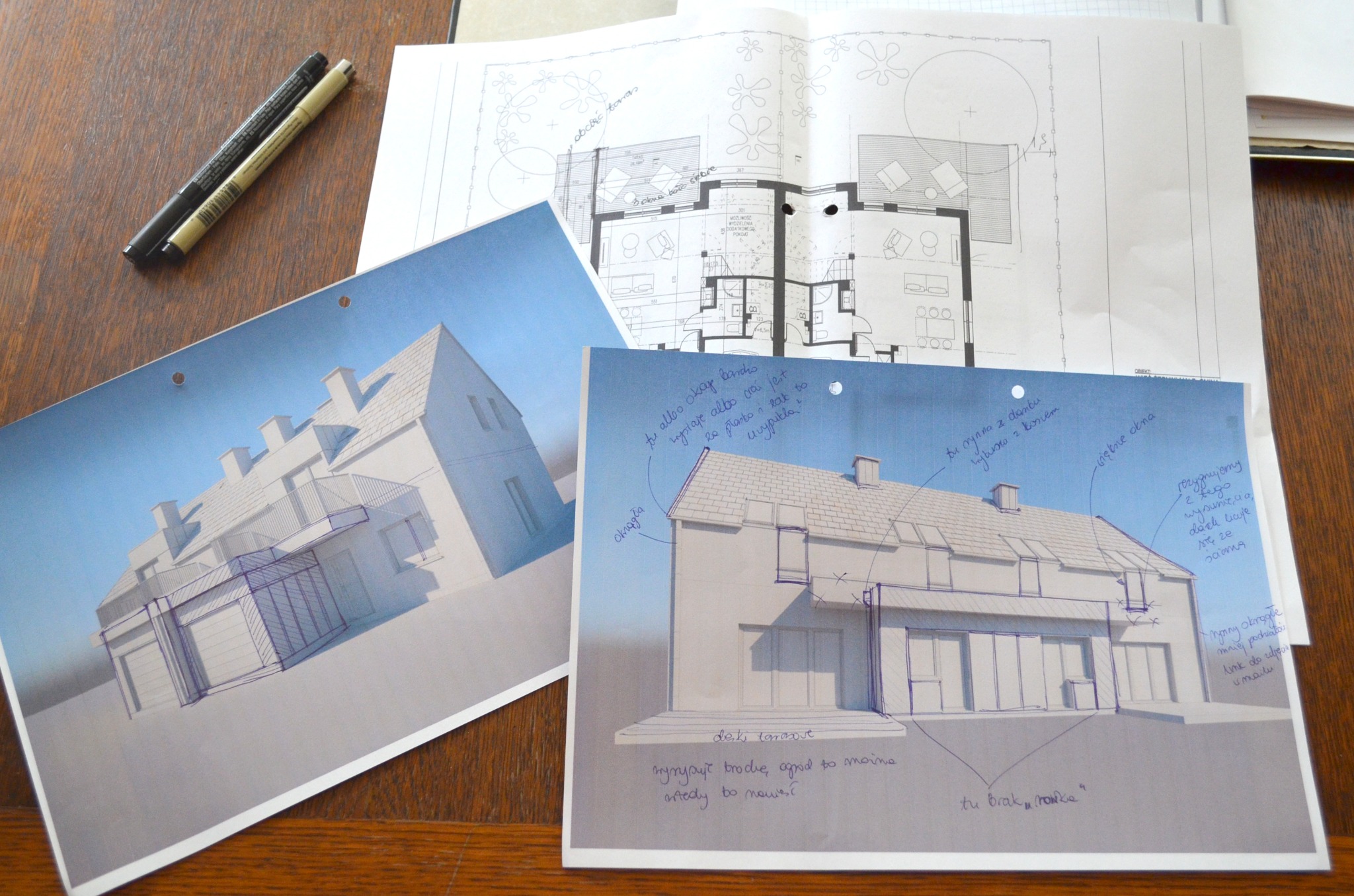 Szkice koncepcyjne domu jednorodzinnego z naniesionymi odręcznymi notatkami, widoczne rzuty i wizualizacje, leżące na drewnianym stole obok dwóch pisaków.