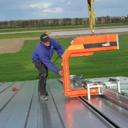 Pracownik w niebieskiej bluzie obsługuje pomarańczowe urządzenie podwieszone na haku dźwigu, układające metalowe płyty na dachu hali; w tle zielone pola i błękitne niebo.