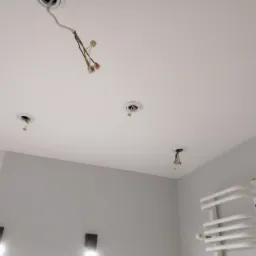 Biały sufit z widocznymi otworami na oświetlenie, z których wystają przewody z kostkami elektrycznymi, na szarej ścianie widoczny grzejnik drabinkowy i otwór w ścianie.