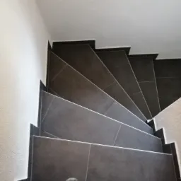 Klatka schodowa wykończona ciemnoszarą, matową glazurą, widok z góry na stopnie i fragment butów osoby stojącej na schodach.